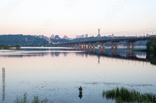 Morning panorama of Dnipro river with Paton Bridge and one fisher. View of Kyiv's hills, Motherland Monument on horizon. Summer in Kyiv, Ukraine. photo