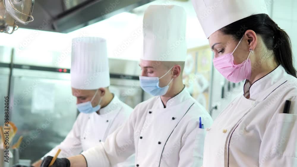Chefs in protective masks and gloves prepare food in the kitchen of a restaurant or hotel Chefs in protective masks and gloves prepare food in the kitchen of a restaurant or hotel