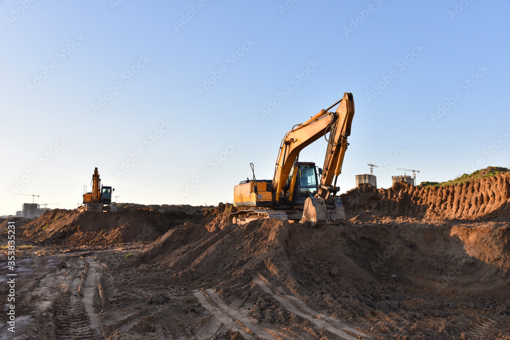 Excavators working at construction site on earthworks. Backhoe digging building foundation. Paving out sewer line. Construction machinery for excavating, loading, lifting and hauling of cargo Excavators working at construction site on earthworks. Backhoe digging building foundation. Paving out sewer line. Construction machinery for excavating, loading, lifting and hauling of cargo