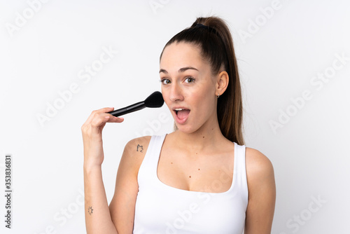 Young brunette woman over isolated white background holding makeup brush and surprised