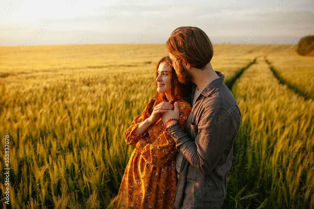 young happy couple in a barley field at sunset. girl in a yellow dress and a guy with a beard. 
portrait of couple walking in the grass