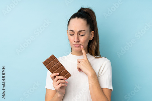 Young brunette woman over isolated blue background taking a chocolate tablet and having doubts