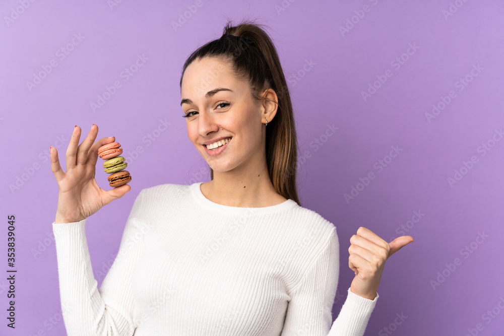 Young brunette woman over isolated purple background holding colorful French macarons and pointing side Young brunette woman over isolated purple background holding colorful French macarons and pointing side
