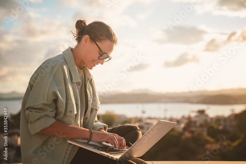 Obraz na płótnie Beautiful young girl woman in eyeglasses sitting with a laptop on her balcony at sunset with a view of the city, remote work from home, a successful freelancer w salonie