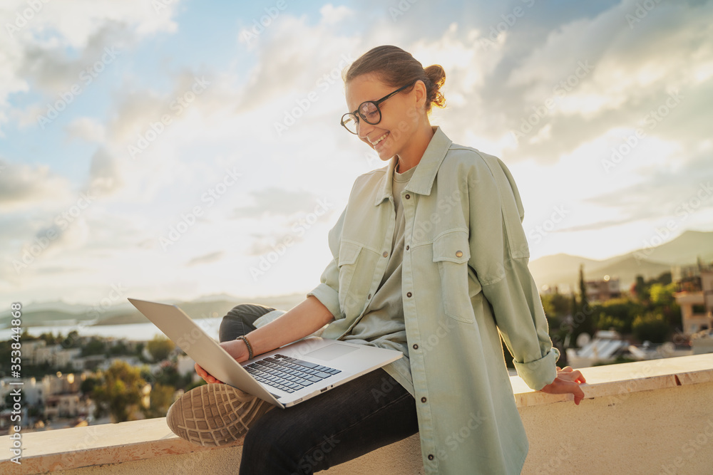 Beautiful young girl woman in eyeglasses sitting with a laptop on her balcony at sunset with a view of the city, remote work from home, a successful freelancer