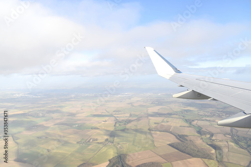 High angle view of landscrape of green land under white airplane wing with clouds in blue sky. View from airplane window