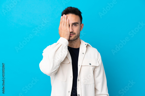 Young handsome man over isolated blue background covering a eye by hand