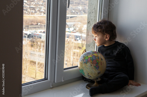 cute curly kid with a globe in his hands looks out the window into the street sitting on the windowsill during quarantine 