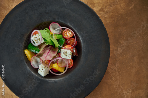 Classic greek salad with red onion, feta cheese, cucumber, bell pepper in a black plate on a copper, metal background. Close up view.