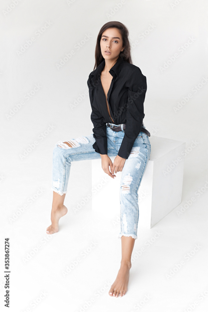 Young cool hipster girl in black shirt and blue banana jeans girl sitting on white cube on white background Young cool hipster girl in black shirt and blue banana jeans girl sitting on white cube on white background