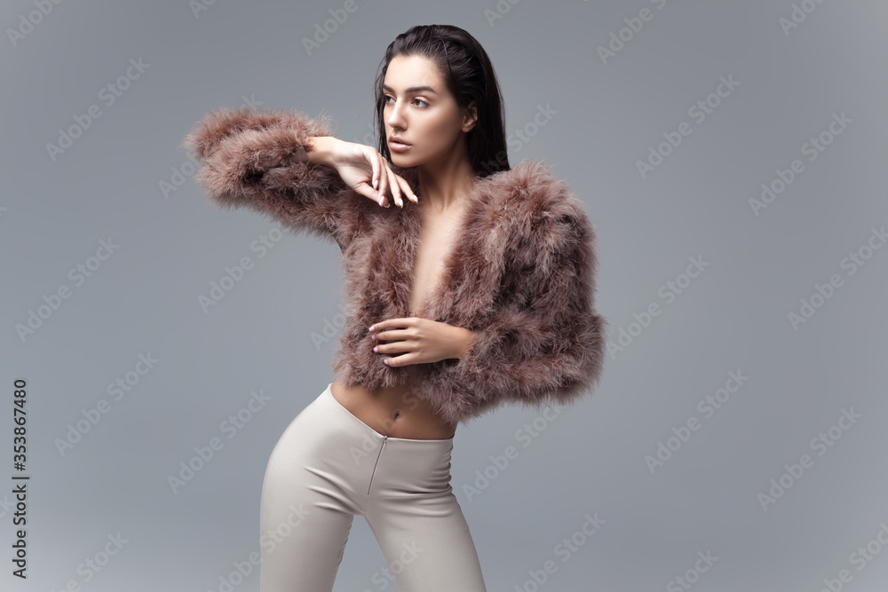Young elegant girl posing in fluffy pink cape with artificial fur and beige flare trousers on gray background Young elegant girl posing in fluffy pink cape with artificial fur and beige flare trousers on gray background