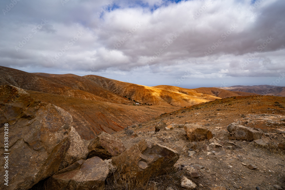 Hügelkette in einer Wüstenlandschaft mit Sonnenflecken und bewölktem Himmel darüber