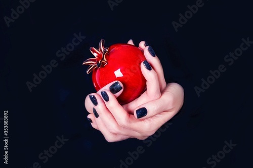 Female hands holding red vase in the shape of a pomegranate fruit on a black background. Manicure with black nail polish. Copy space