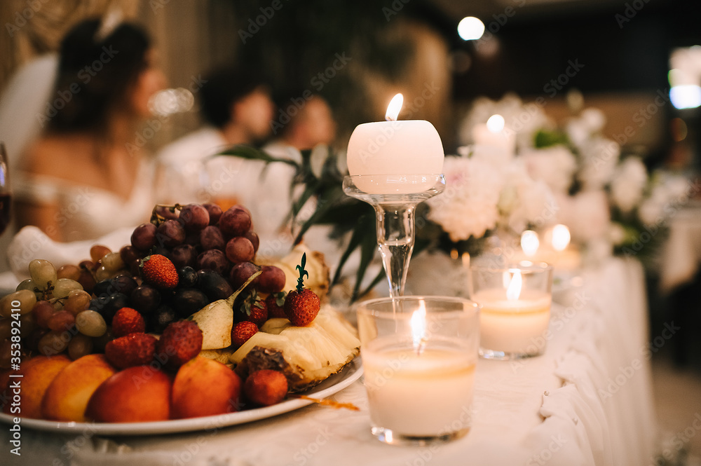 Fototapeta premium Fruits on a plates, Grapes, pineapple, strawberries, peach, plum. Reception, catering. The table is covered with a white tablecloth. Candles are burning