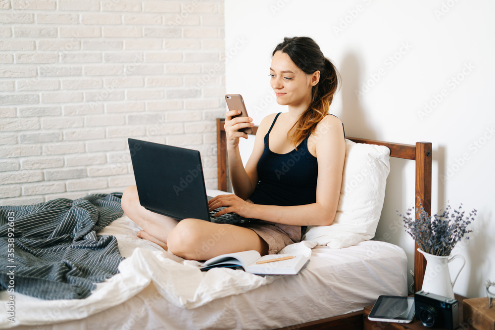 young European woman using a laptop works remotely from home, in her bed, talking on smartphone with a client, while the virus is quarantined.Cozy workplace, e-learning concept.freelancer young European woman using a laptop works remotely from home, in her bed, talking on smartphone with a client, while the virus is quarantined.Cozy workplace, e-learning concept.freelancer