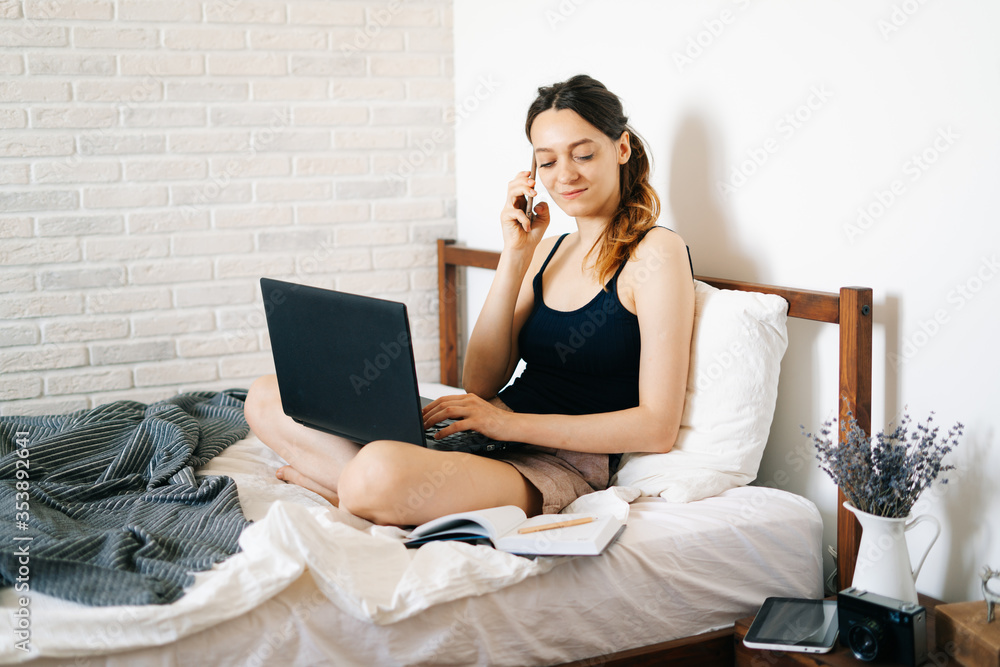 young European woman using a laptop works remotely from home, in her bed, talking on smartphone with a client, while the virus is quarantined.Cozy workplace, e-learning concept.freelancer young European woman using a laptop works remotely from home, in her bed, talking on smartphone with a client, while the virus is quarantined.Cozy workplace, e-learning concept.freelancer