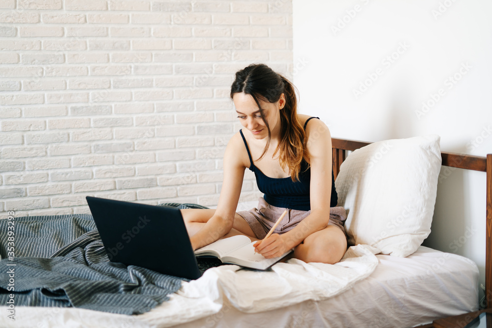 European woman, using a laptop, studying information on the Internet in her bed, writes notes in her diary,while the virus is quarantined.Cozy workplace, e-learning concept.freelancer European woman, using a laptop, studying information on the Internet in her bed, writes notes in her diary,while the virus is quarantined.Cozy workplace, e-learning concept.freelancer