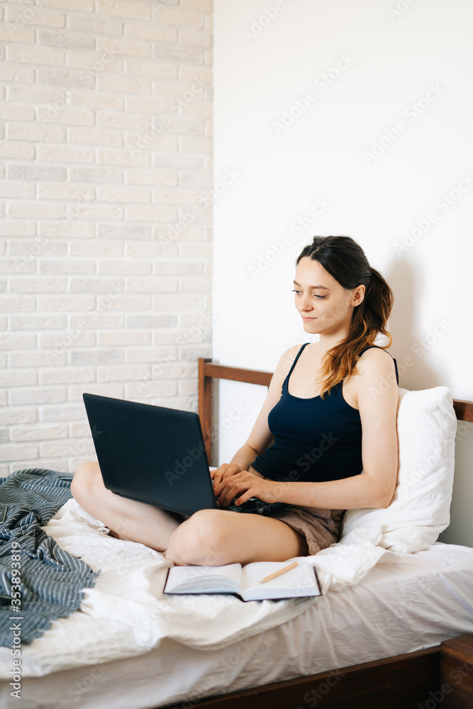 A young woman, using a laptop, communicates over the Internet with a client in her bed, while the virus is quarantined.Cozy office workplace,Remote work, e-learning concept.freelancer A young woman, using a laptop, communicates over the Internet with a client in her bed, while the virus is quarantined.Cozy office workplace,Remote work, e-learning concept.freelancer