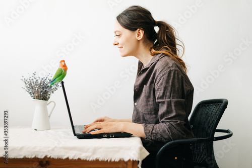 young European woman, working from home during the virus quarantine, on her laptop.a small green lovebird parrot sits on a laptop monitor.A cosy working place,the concept of remote work. freelance