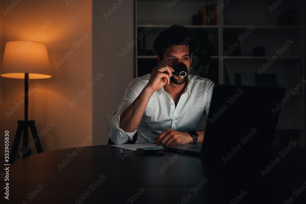 Young businessman drinking coffe and using a laptop computer at his desk in the office or home late into the night Young businessman drinking coffe and using a laptop computer at his desk in the office or home late into the night