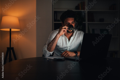 Young businessman drinking coffe and using a laptop computer at his desk in the office or home late into the night