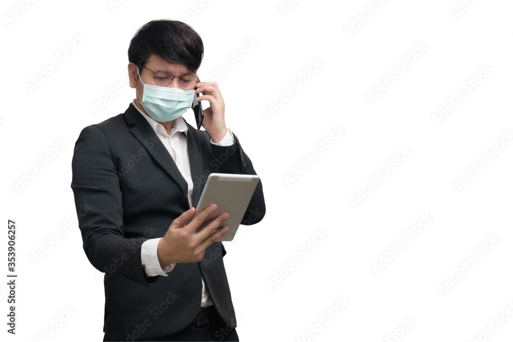 Asian businessman wearing surgical face mask in formal black suit jacket, using smartphone to talking and look at the tablet, studio lighting isolate on white background, coronavirus, COVID19 concept