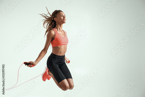 Flying. Happy and young african woman with perfect body skipping rope while exercising in studio against grey background
