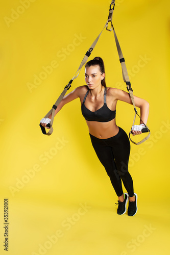 Working hard. Full length of young athletic woman in sportswear doing push ups with trx fitness straps against yellow background. Studio shot photo