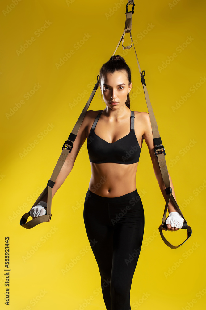 TRX exercises. Young sporty woman in sportswear doing push ups with trx fitness straps against yellow background. She is looking at camera. Studio shot TRX exercises. Young sporty woman in sportswear doing push ups with trx fitness straps against yellow background. She is looking at camera. Studio shot