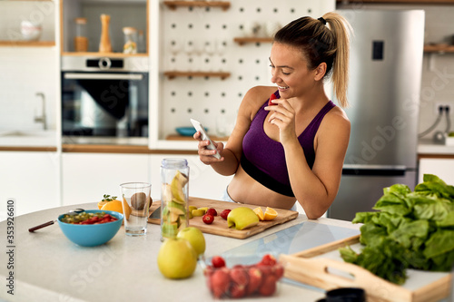 Happy sportswoman texting on cell phone while eating fruit in the kitchen.
