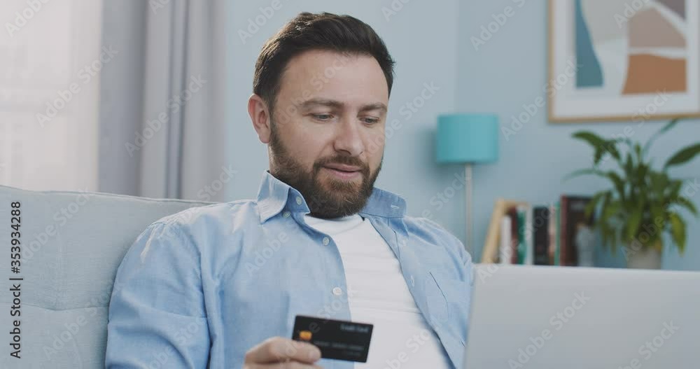 Close up portrait concentrated man holding credit card, writing payment document, using laptop for online shopping while sitting at sofa. Male doing financial transaction with credit card and smiling. Close up portrait concentrated man holding credit card, writing payment document, using laptop for online shopping while sitting at sofa. Male doing financial transaction with credit card and smiling.