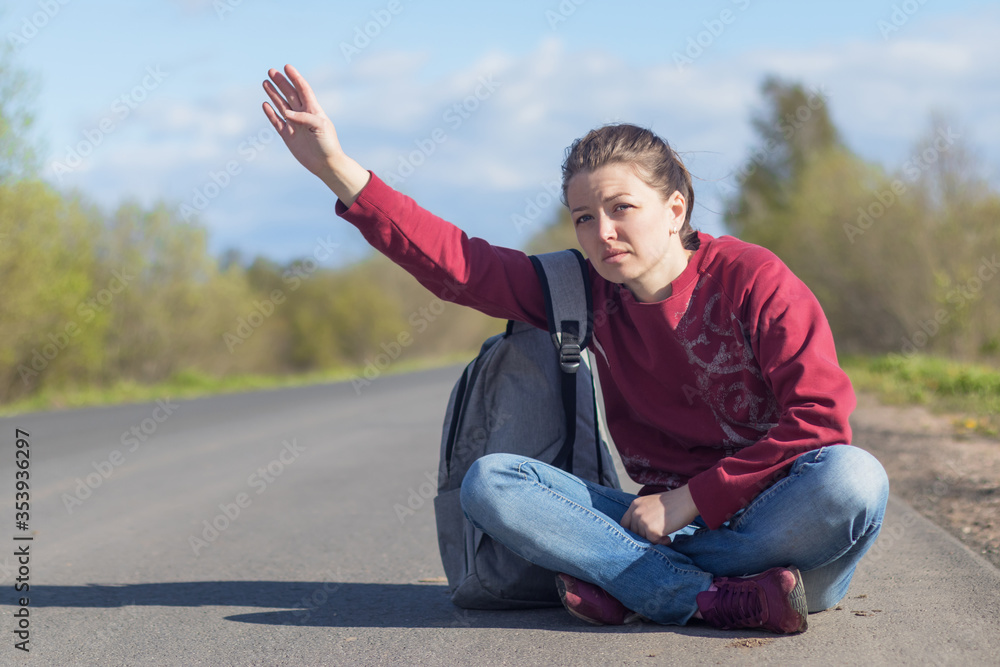 passenger. tired girl is sitting on the road on roadside with backpack and hand up gesture stops the car. hitchhiking young woman.