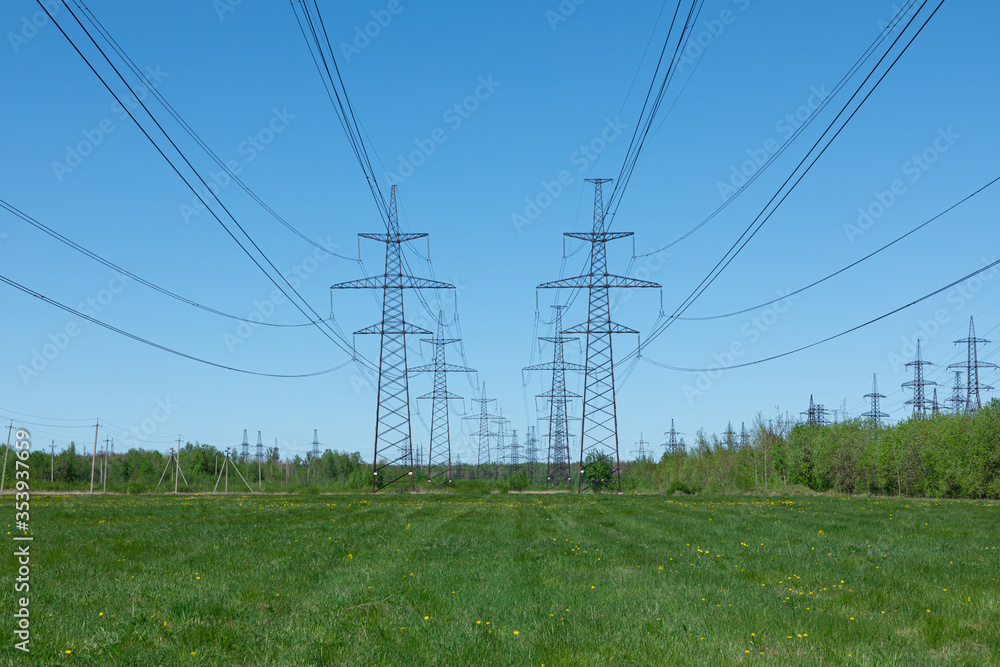 The towers of electric main in the countryside field on the background of blue sky and the forest with the wires in sunny summer or spring weather The towers of electric main in the countryside field on the background of blue sky and the forest with the wires in sunny summer or spring weather