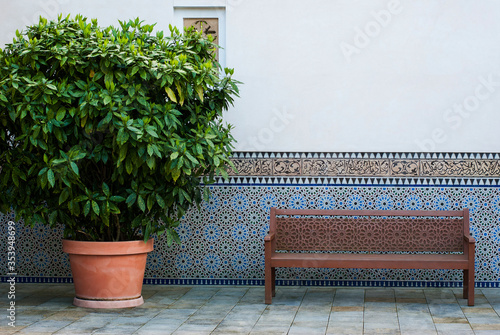 Fototapeta Naklejka Na Ścianę i Meble -  A wooden bench with patterns stands by the wall which is lined with blue tiles, a large green plant in the pot on the left. The courtyard is decorated in oriental style.