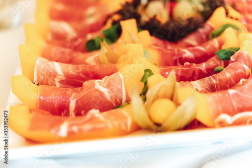 Closeup of tray with juicy slices of yellow melon rolled into ham. Self-service buffet table. Celebration, birthday, party, wedding concept.