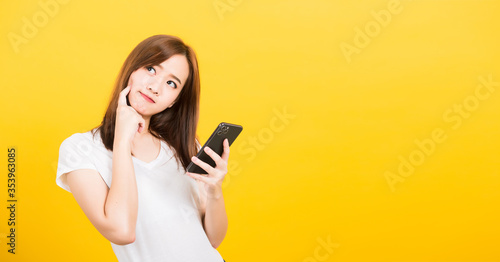 Asian happy portrait beautiful cute young woman teen standing wear t-shirt her using holding smart mobile phone and thinking looking to side isolated, studio shot yellow background with copy space