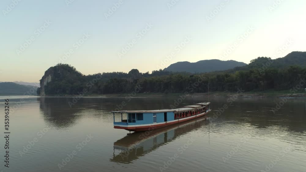 Aerial shot of tourists in passenger boat on river against sky, drone flying over water with reflection near mountains during sunset - Mekong River, Laos Aerial shot of tourists in passenger boat on river against sky, drone flying over water with reflection near mountains during sunset - Mekong River, Laos
