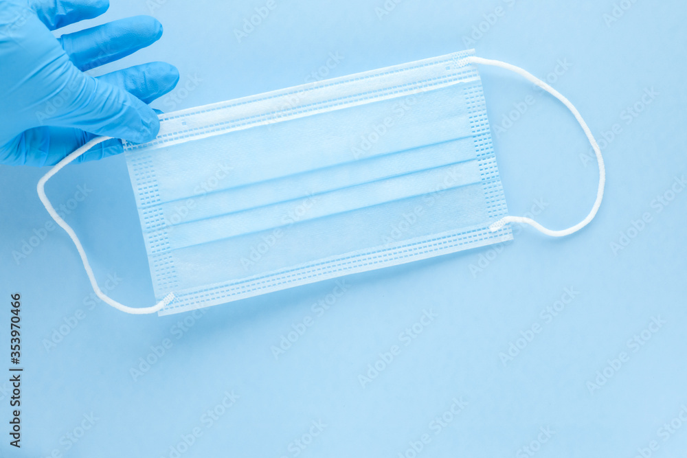 Gloved hand holding a single disposable medical face mask on blue background. Personal protective equipment, PPE.