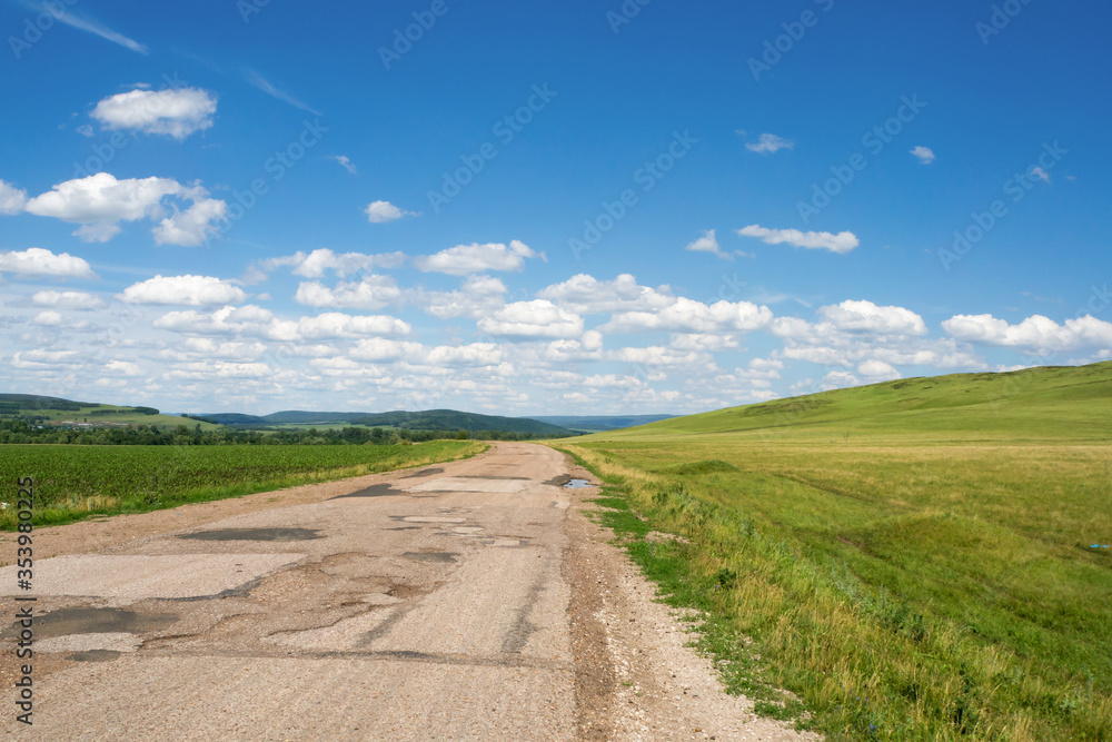 Road with broken worn asphalt pavement. Summer countryside landscape.