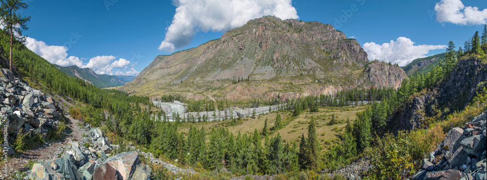 Panoramic view of a mountain valley with a river. Forest, blue sky and hillside trail. Summer landscape.
