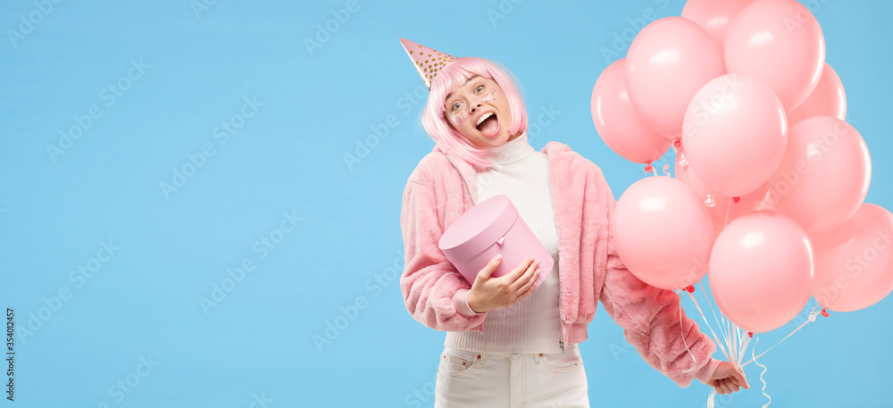Horizontal banner of young birthday girl dressed in pink, holding balloons and gift box, shouting with surprise, isoalted on blue background, copy space on left Horizontal banner of young birthday girl dressed in pink, holding balloons and gift box, shouting with surprise, isoalted on blue background, copy space on left
