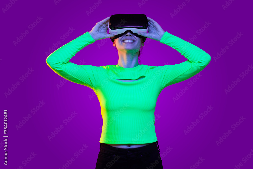 Young smiling teenage girl wearing green neon top, having virtual reality headset on, exploring new gaming opportunities, isolated on purple background Young smiling teenage girl wearing green neon top, having virtual reality headset on, exploring new gaming opportunities, isolated on purple background