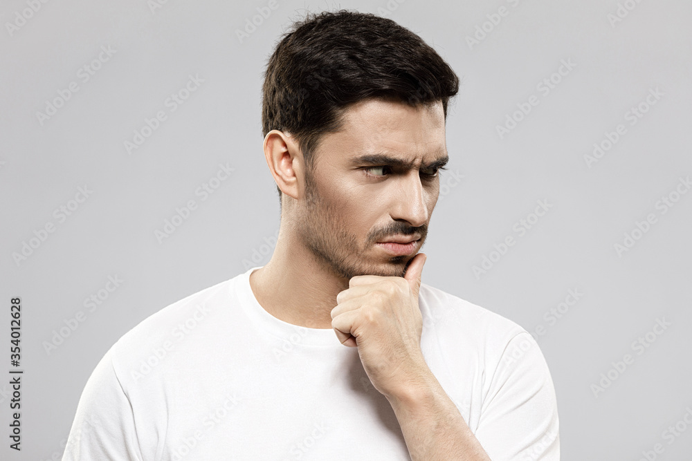 Close-up of young handsome man in white t-shirt feeling doubt while making difficult decision, holding chin and looking aside, isolated on gray background Close-up of young handsome man in white t-shirt feeling doubt while making difficult decision, holding chin and looking aside, isolated on gray background