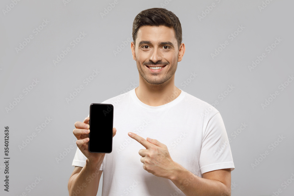 Young handsome man in white t-shirt holding smartphone, pointing to blank screen with finger, recommending mobile application, isolated on gray background with copy space Young handsome man in white t-shirt holding smartphone, pointing to blank screen with finger, recommending mobile application, isolated on gray background with copy space