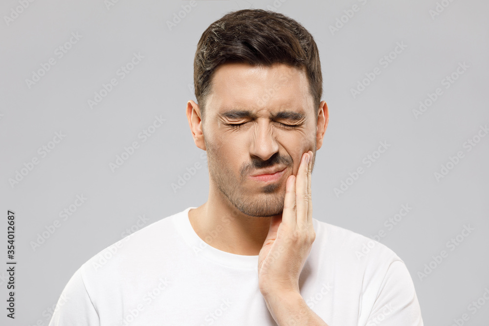 Young man in white t-shirt suffering from severe toothache, touching cheek with fingers, eyes closed because of strong pain, isolated on gray background