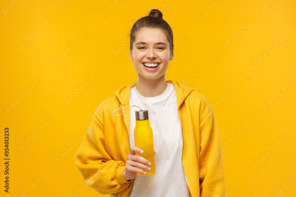 Young sportswoman in bright hoodie and white t-shirt, holding reusable eco water bottle, having fun and feeling good for walk, isolated on yellow background Young sportswoman in bright hoodie and white t-shirt, holding reusable eco water bottle, having fun and feeling good for walk, isolated on yellow background