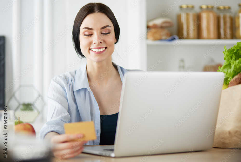 Smiling woman online shopping using computer and credit card in kitchen Smiling woman online shopping using computer and credit card in kitchen