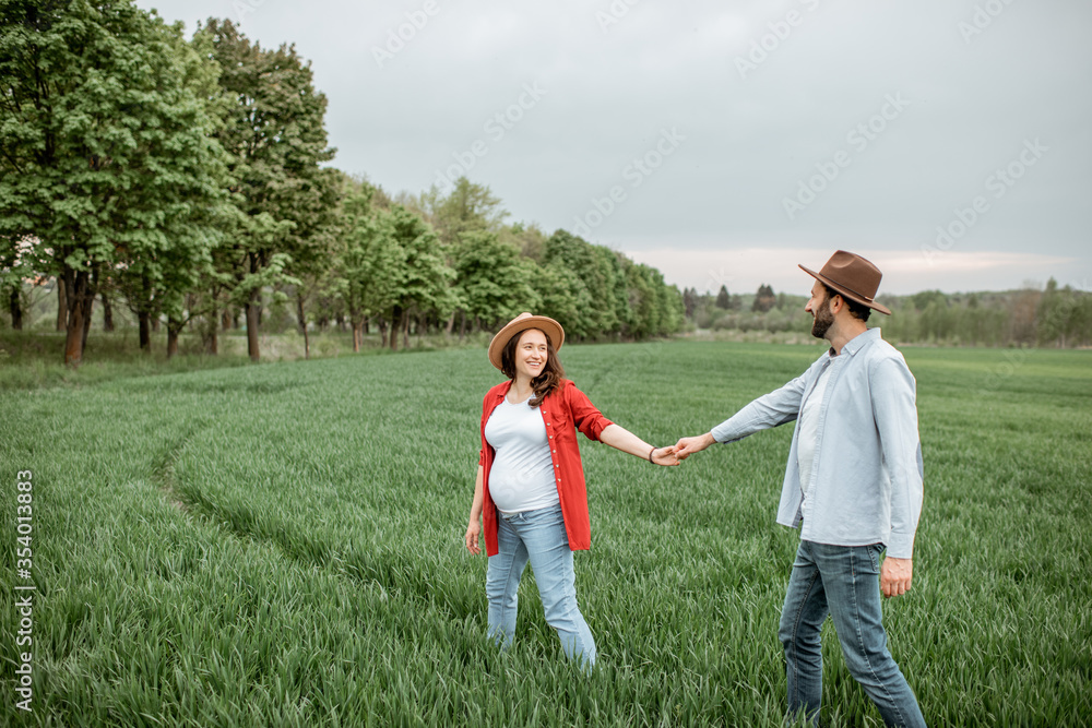 Fototapeta premium Pregnant woman with her man having fun together, walking on the greenfield. Happy couple expecting a baby, young family concept