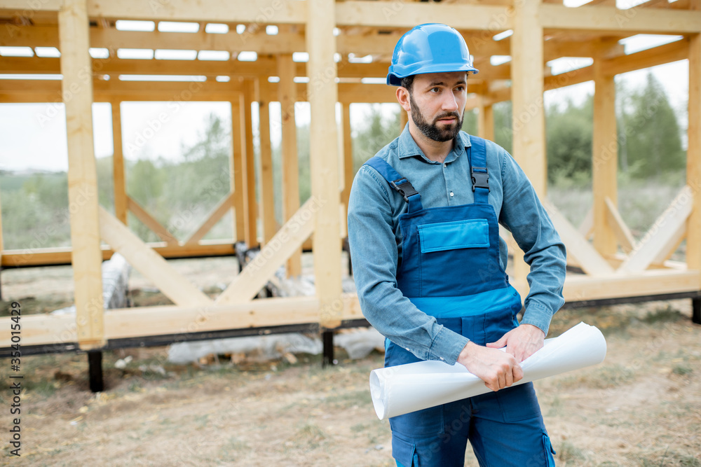 Builder in blue overalls and hard hat with blueprints on the construction site, building wooden frame house Builder in blue overalls and hard hat with blueprints on the construction site, building wooden frame house