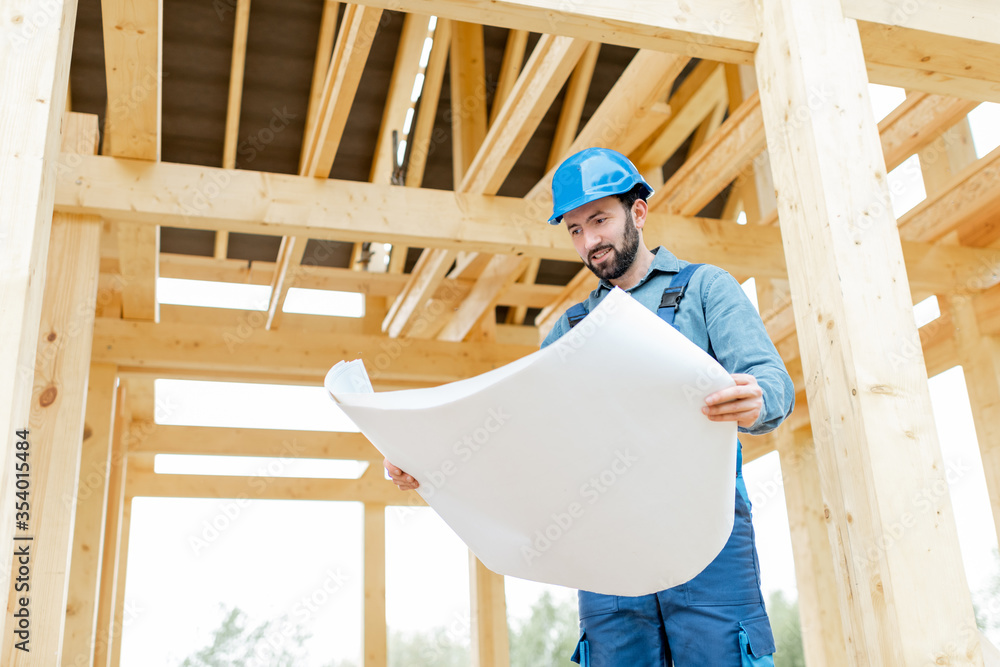Builder in blue overalls and hard hat with blueprints on the construction site, building wooden frame house Builder in blue overalls and hard hat with blueprints on the construction site, building wooden frame house